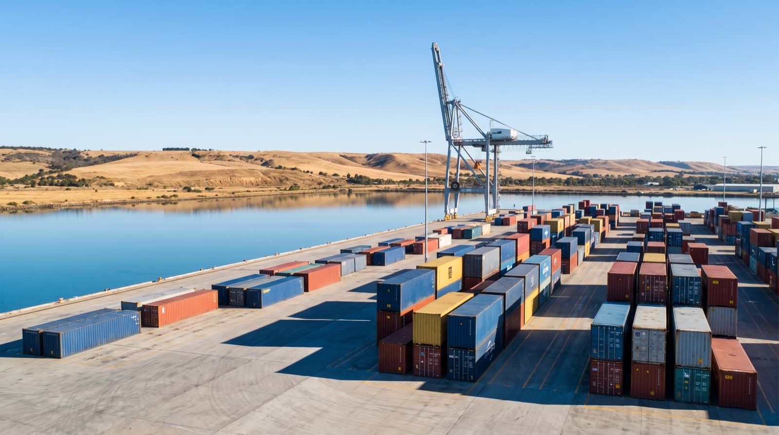 Container wharf at Port Adelaide with shipping containers stacked beside calm harbour water and a single gantry crane
