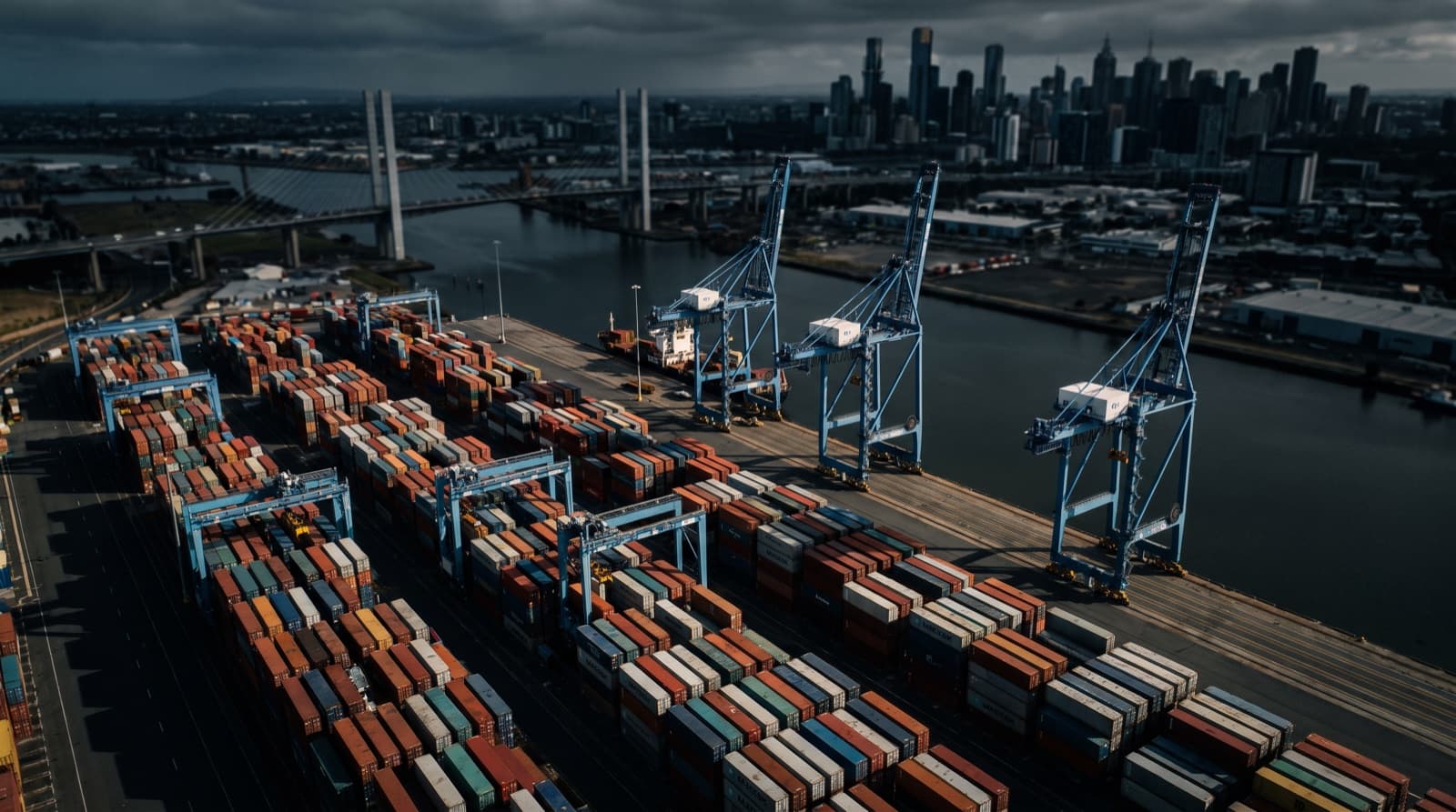 Aerial view of the Port of Melbourne container terminal with blue gantry cranes and the Melbourne skyline in the background