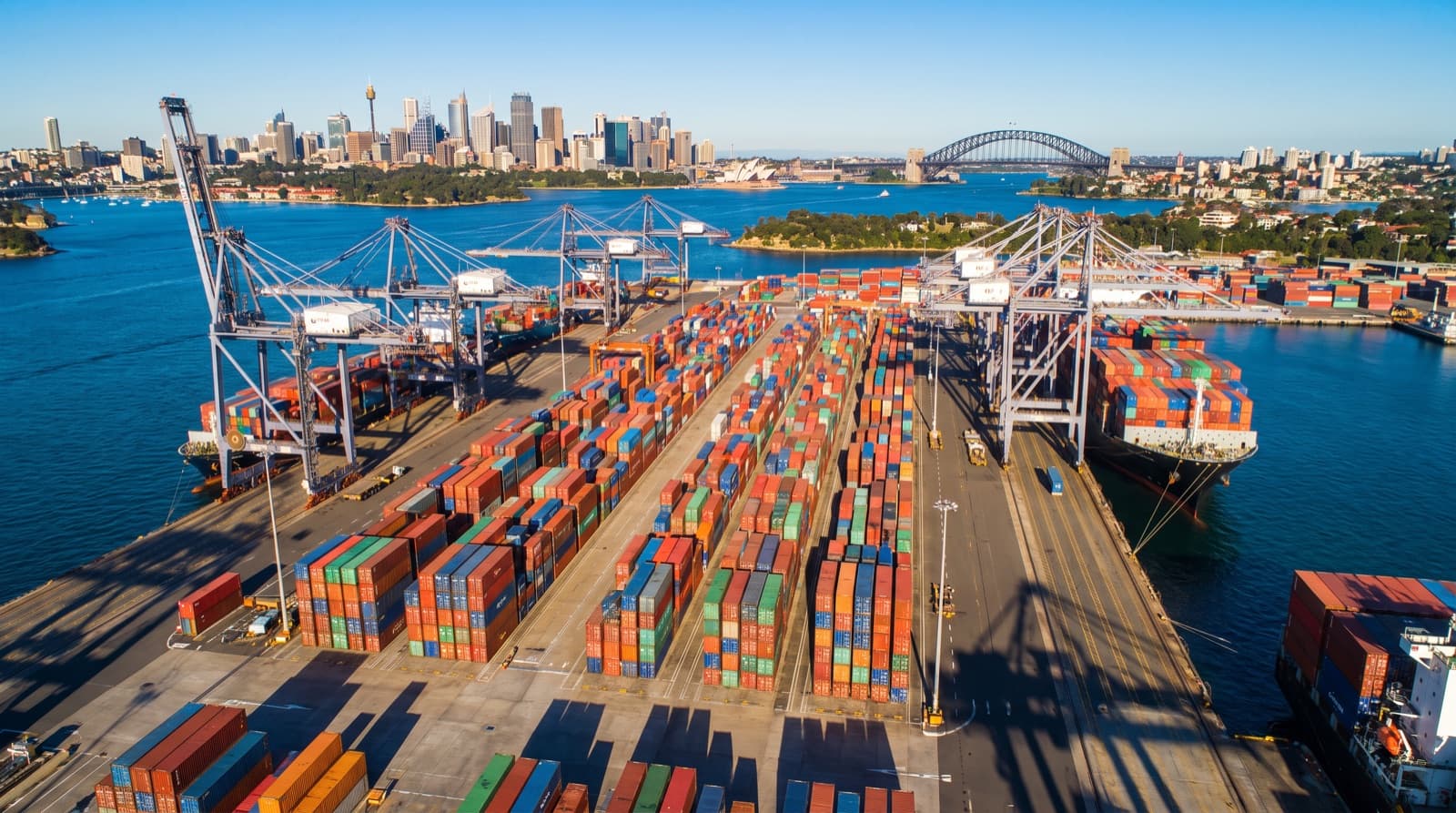 Aerial view of a container shipping terminal in Sydney with stacked containers, gantry cranes and the city skyline beyond